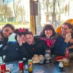 Gruppo di sei persone sedute al tavolo durante il Carnevale 2026 di ELO Epilessia Lombardia ETS, alcuni in maschera e altri senza, che si abbracciano